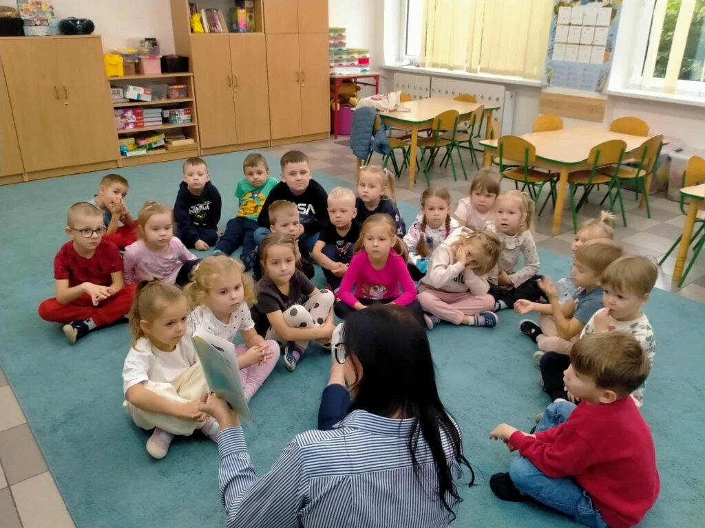 dzieci siedza na dywanie z kobietą która trzyma książkę